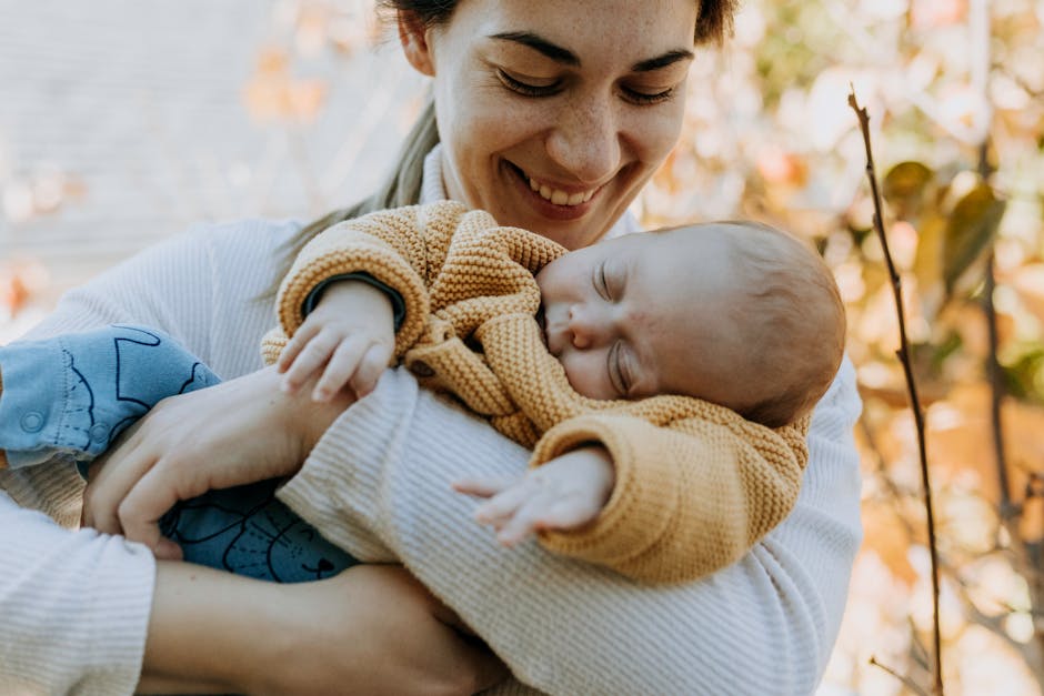 A joyful mother in autumn attire proudly holds her sleeping baby in a peaceful outdoor setting.