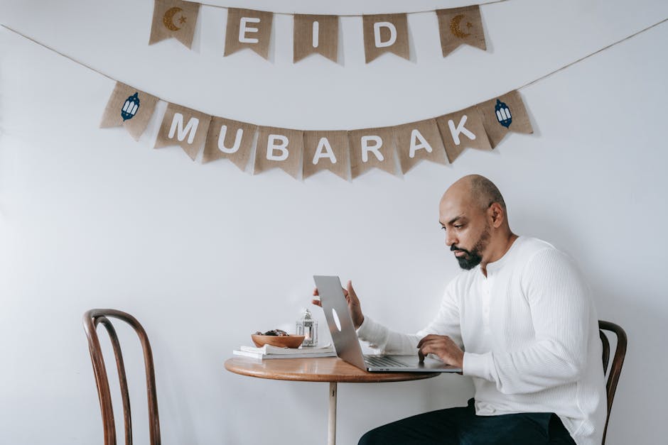 A focused man working on a laptop during Eid Mubarak celebration indoors, blending work and festivity.
