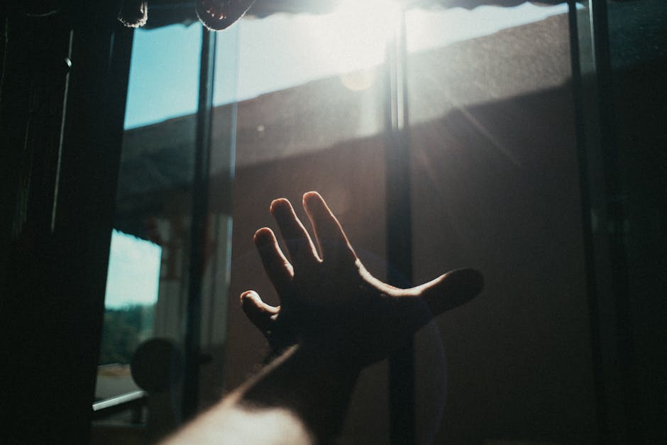 A hand reaches towards the bright sunlight streaming through a window indoors.
