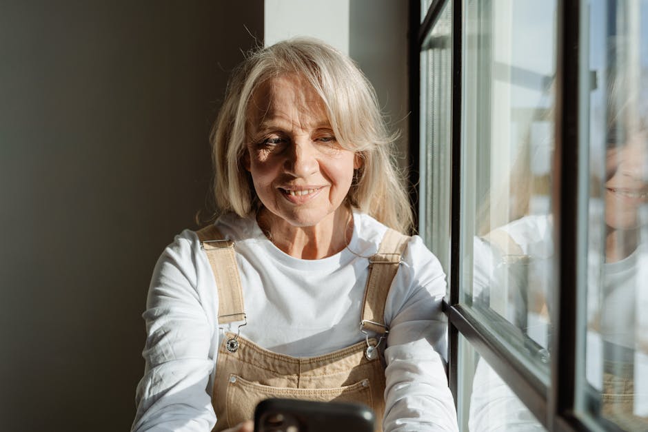 Elderly woman enjoying leisure time using smartphone, bathed in soft natural light by the window.