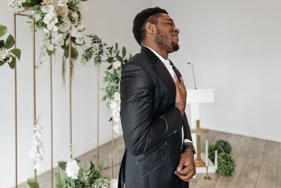 Grateful groom in black tuxedo standing happily at an elegant indoor wedding ceremony with floral decorations.