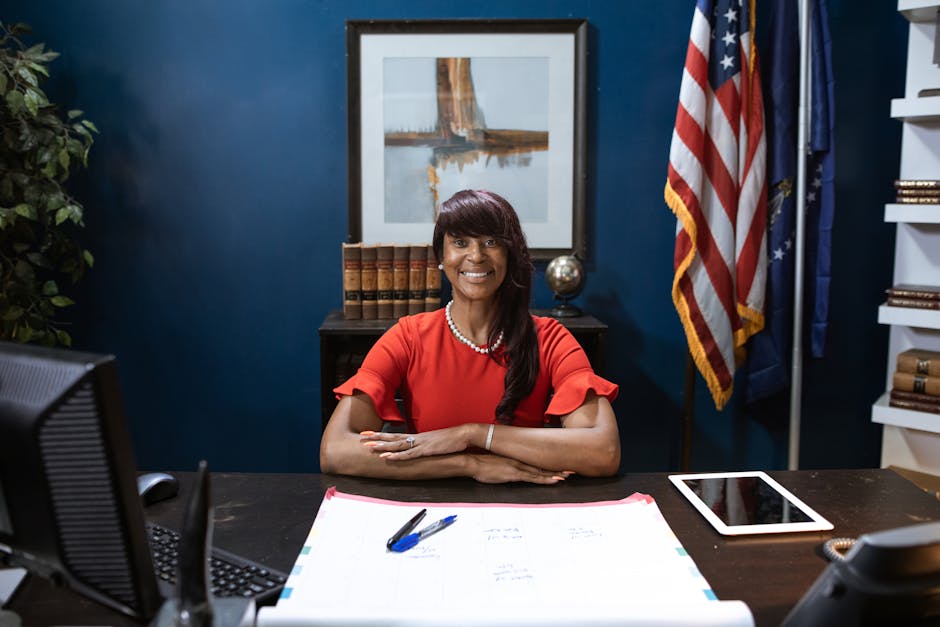A smiling businesswoman sitting at a desk in a professional office with USA flag in the background.