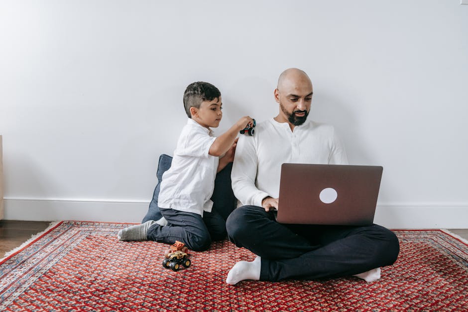 A father and his son enjoying time together with a laptop and toy car on a carpeted floor.