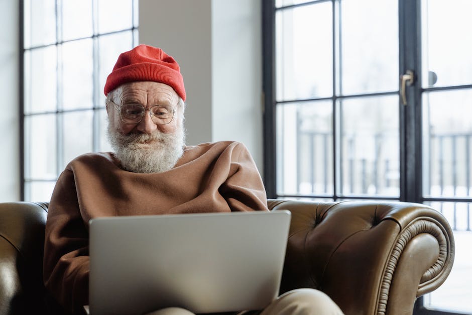 An elderly man with a beard uses a laptop in a cozy indoor setting, smiling contently.