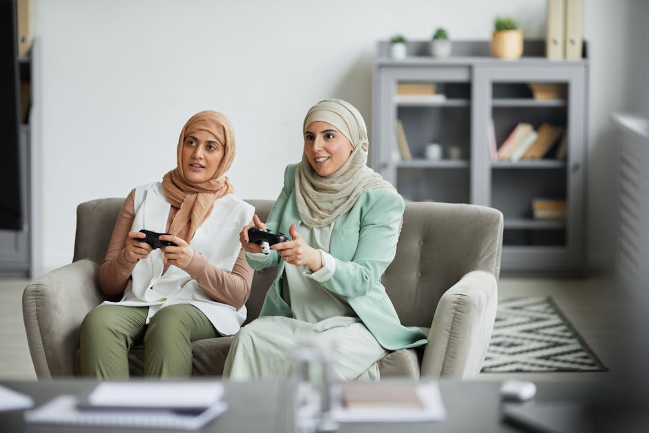 Two Muslim women in hijabs play video games on a sofa, showcasing cultural diversity and lifestyle.