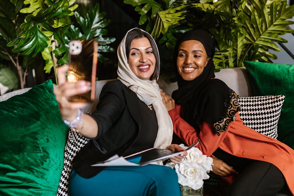 Two smiling women in hijabs take a selfie on a vibrant and stylish couch indoors.