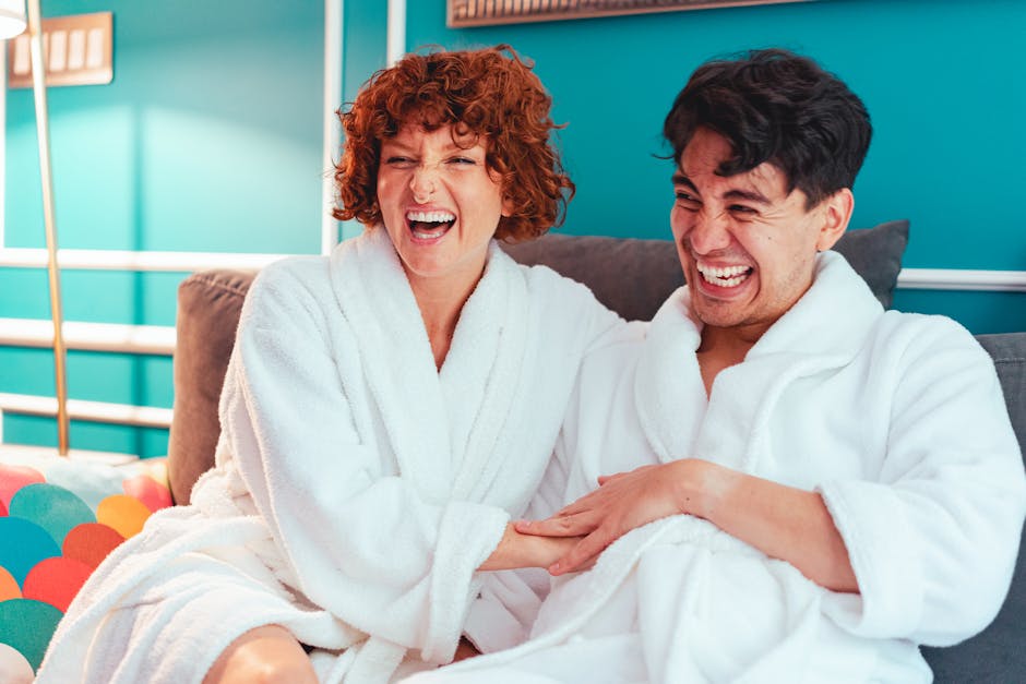 A happy couple laughing together at home, wearing bathrobes on a sofa.