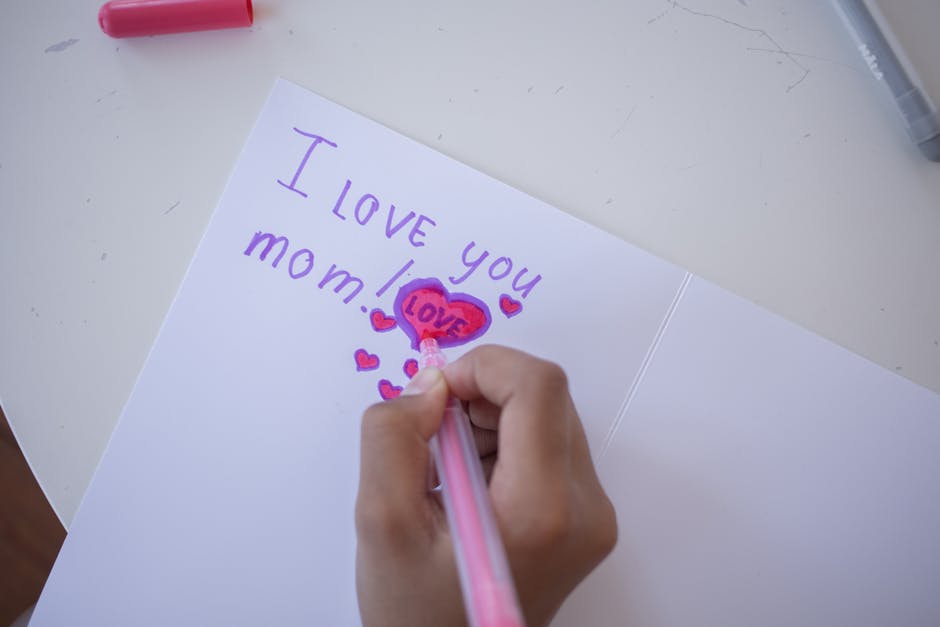 A child drawing 'I love you mom!' in a greeting card with colored pens.