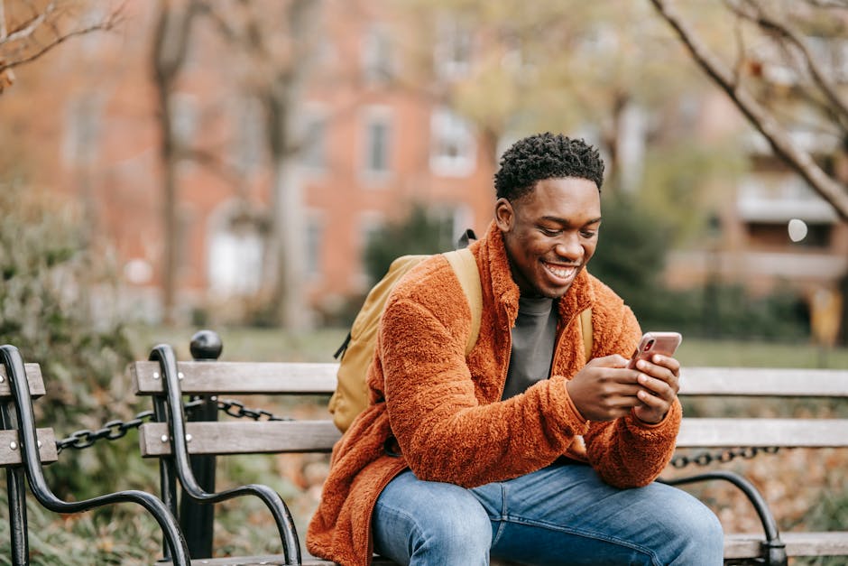 A cheerful young man in warm clothes smiling while using his phone on a bench in a city park.