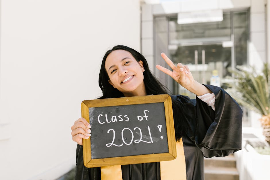 Smiling graduate in cap and gown holding a 'Class of 2021' sign while making a peace sign.