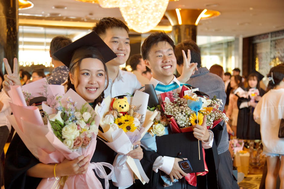 Three students celebrating graduation with flowers and smiles indoors.