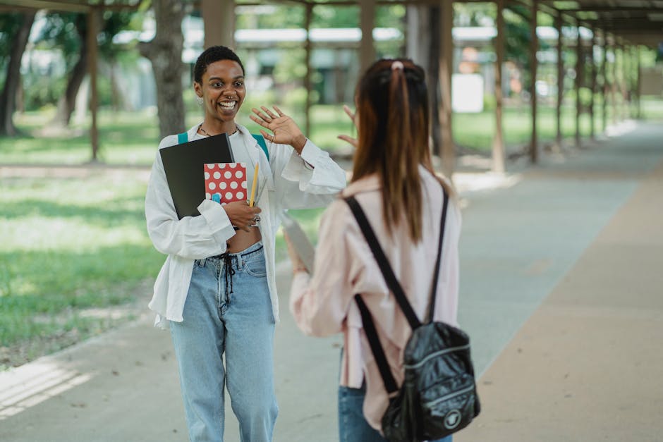 Young cheerful African American woman with workbooks saying hello to unrecognizable friend with rucksack while looking at each other on walkway