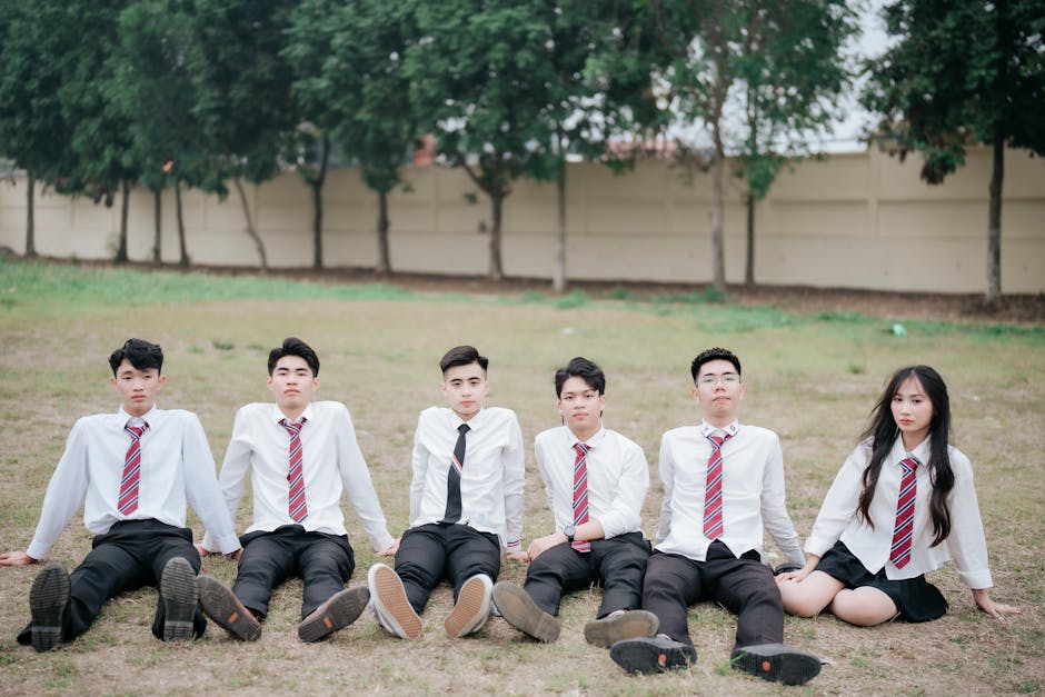 A group of teenagers in school uniforms sitting on grass, outdoors against a tree-lined fence.