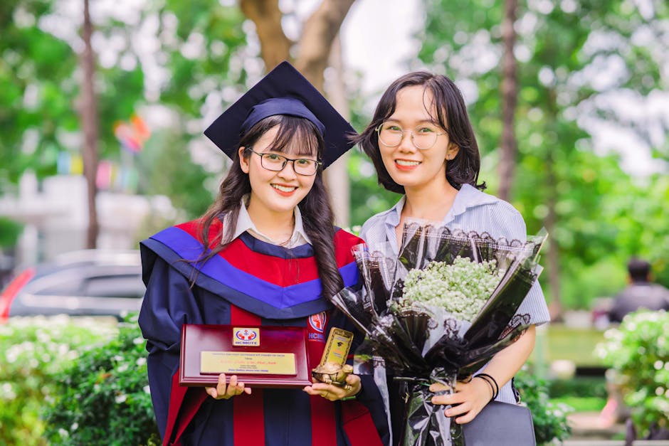 Two women celebrating graduation with smiles, gowns, and bouquets outdoors.