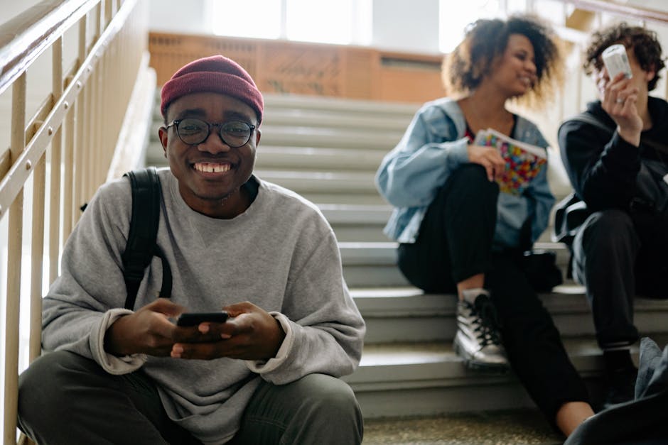 Diverse group of happy students sitting on school staircase, smiling and using mobile phones.