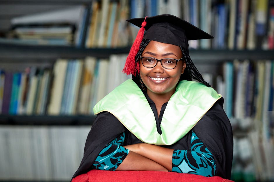 Smiling woman in graduation gown and cap sitting in a library, looking confident.