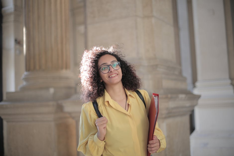 Smiling young woman with curly hair and eyeglasses on campus, holding files.