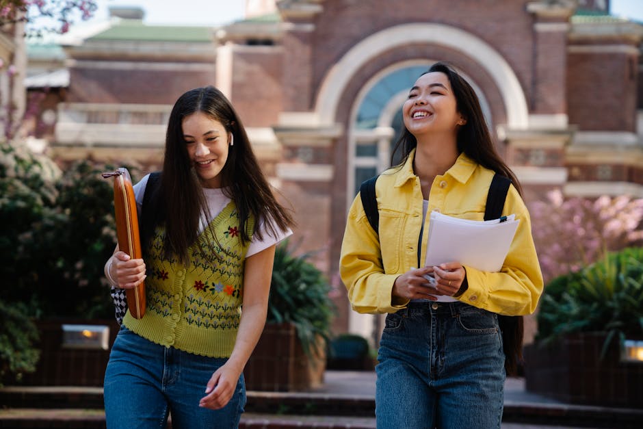 Two smiling young women walking outdoors on a university campus, holding books.