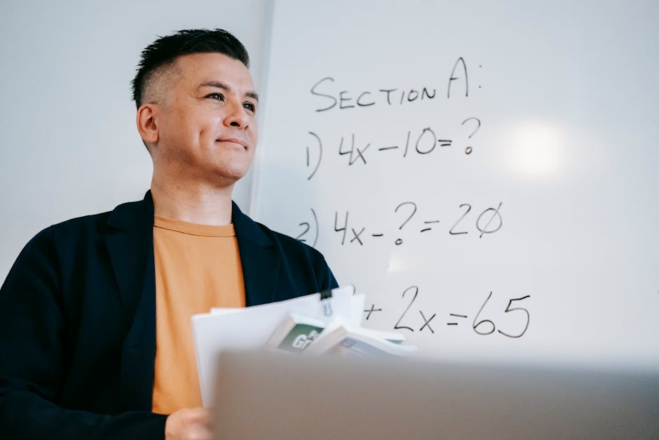 Smiling teacher holding papers, explaining algebra equations on a whiteboard in a classroom setting.