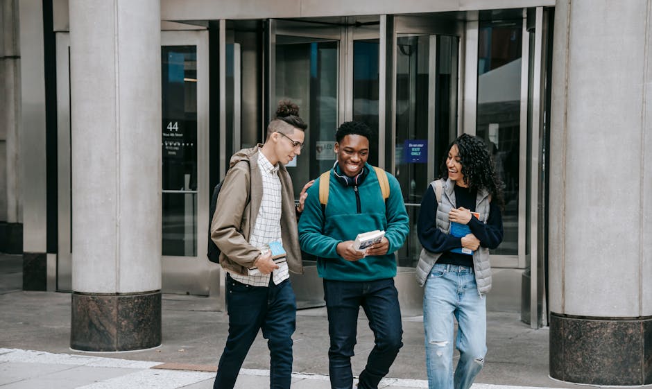 Smiling young multiracial group of friends in casual outfits and backpacks strolling with books and talking on city street near building with columns and glass doors in daylight