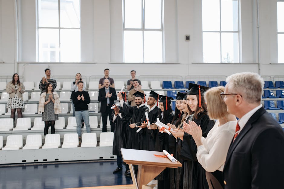 Graduates in gowns receive applause during an indoor ceremony, celebrating achievement.
