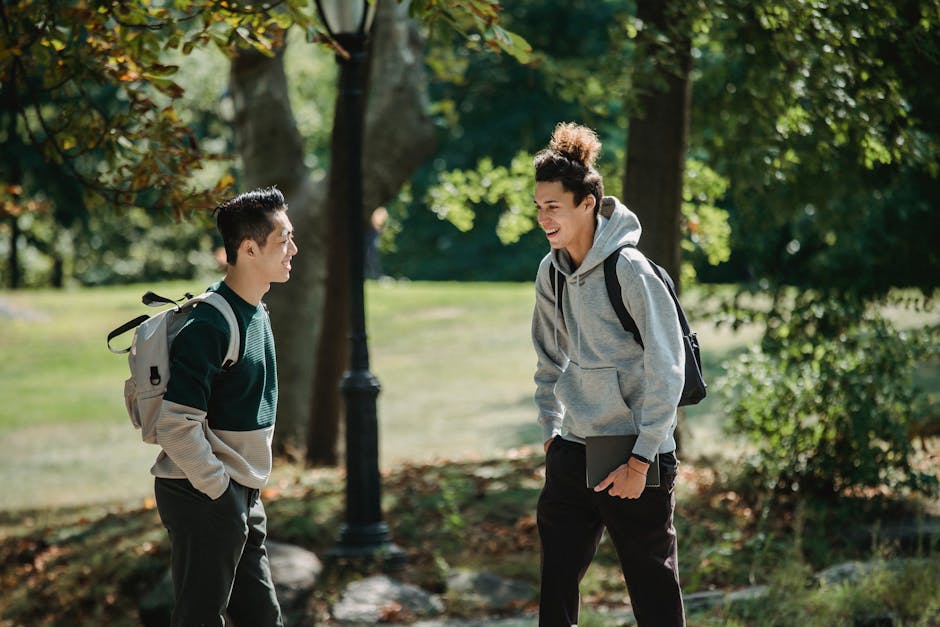 Cheerful diverse male students with backpacks and hands in pockets standing in park and looking at each other