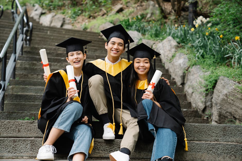 Three happy graduates in caps and gowns celebrate on outdoor steps, holding diplomas.