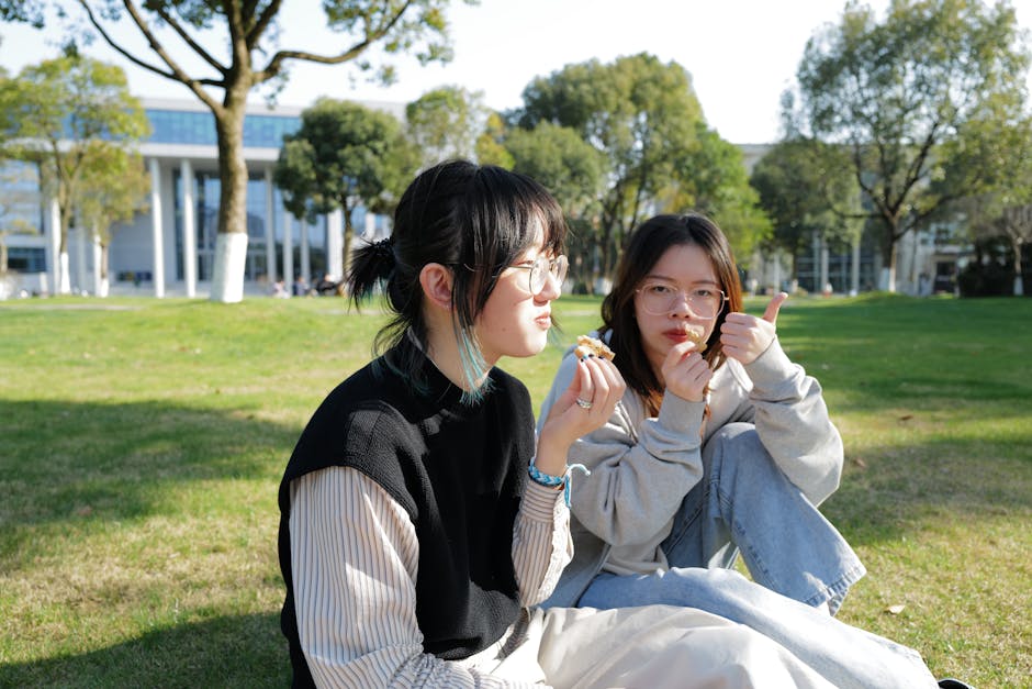 Two friends relaxing on a grassy field, enjoying snacks together on a sunny campus day.