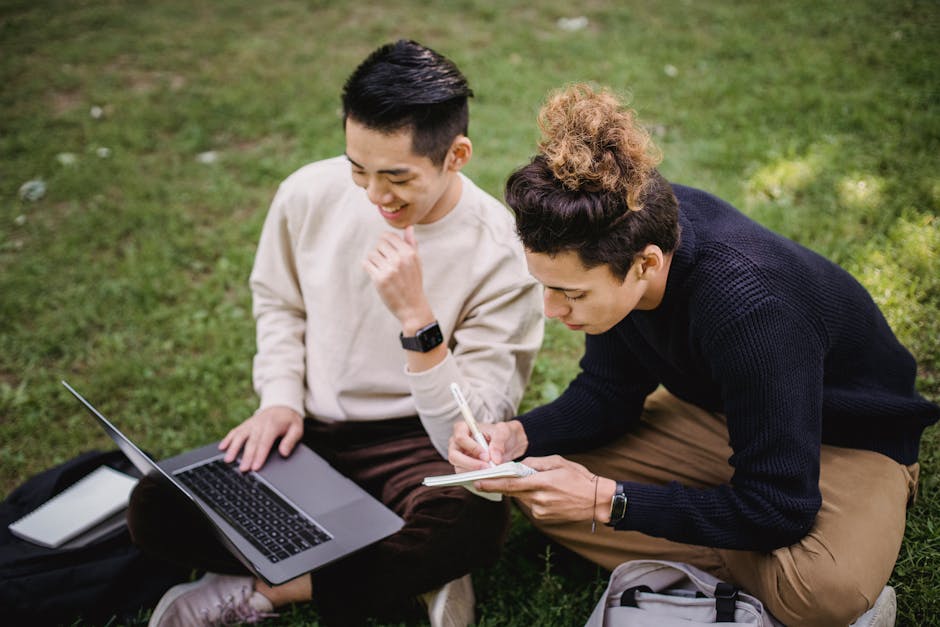 Two young men studying together on a laptop in an outdoor park setting.