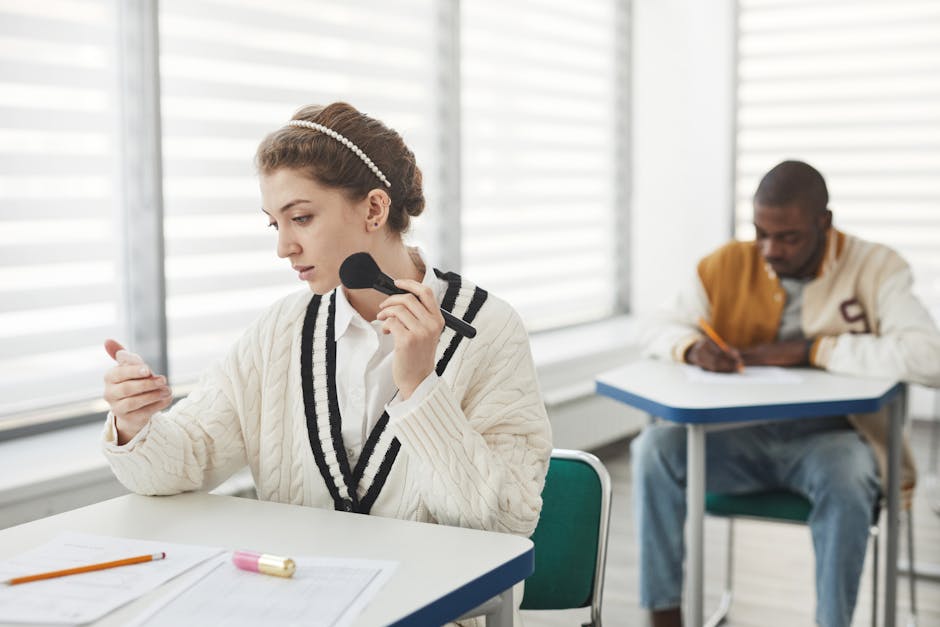 Two diverse students in a classroom, one using makeup, the other taking notes.