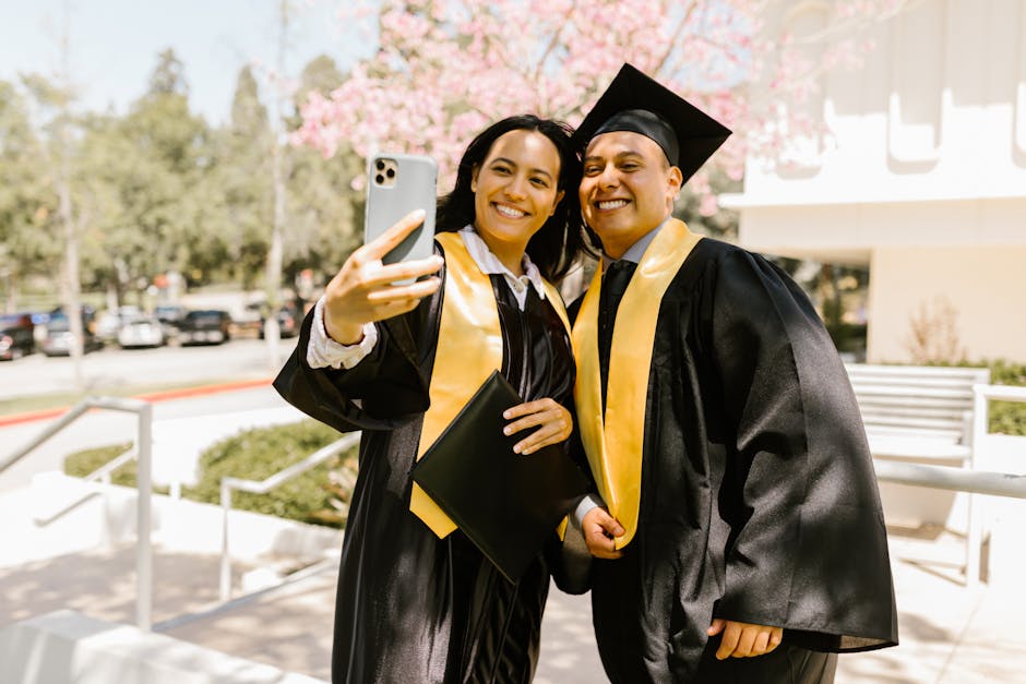 Two graduates in gowns taking a joyful selfie outdoors on a sunny day, celebrating their achievement.