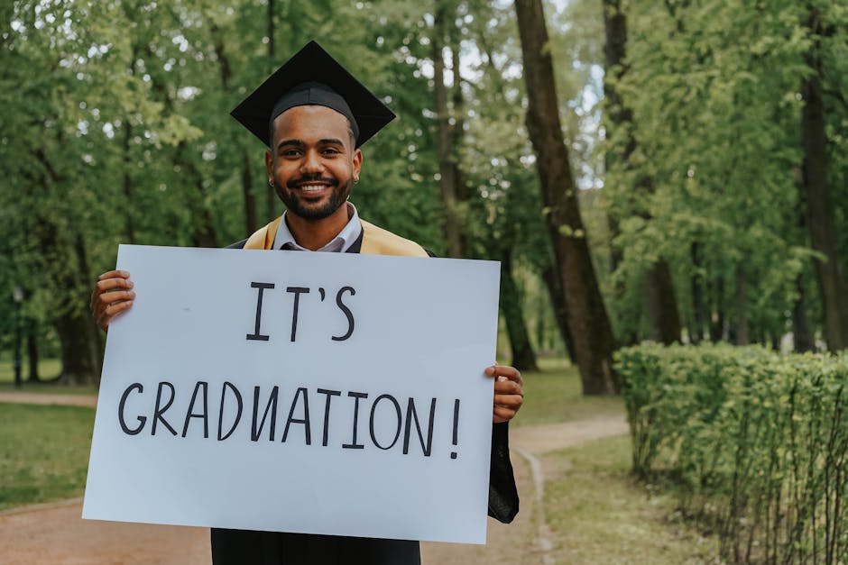 Young graduate smiling while holding a graduation sign in a park.