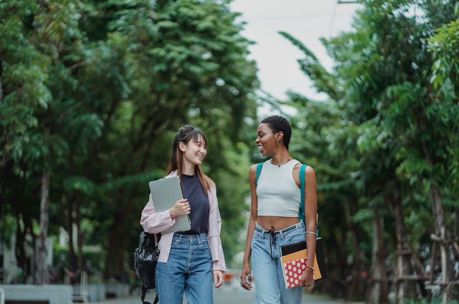 Two women smiling and walking through a tree-lined pathway, carrying books and laptops, enjoying a day outdoors.
