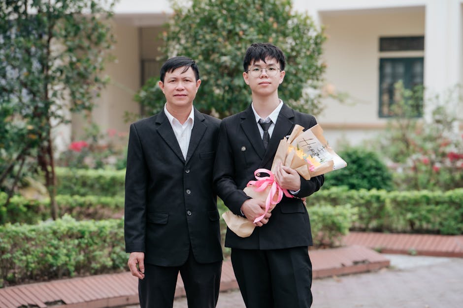A father and son wearing suits celebrating graduation with flowers outdoors.