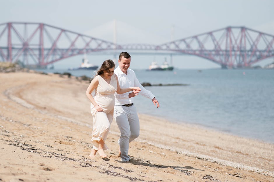Pregnant couple enjoying a relaxed walk on a beach near a scenic bridge, expressing love and joy.
