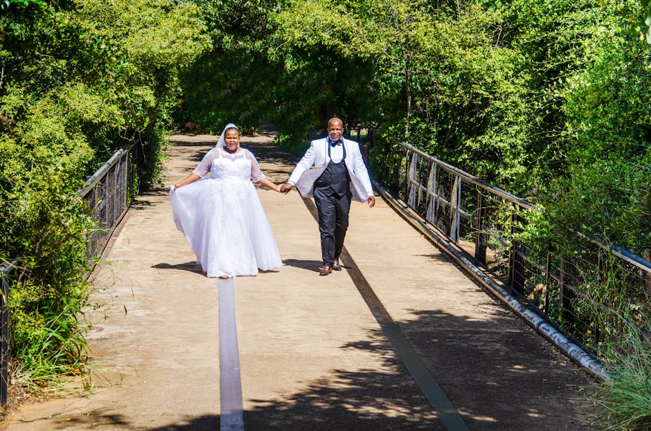 Happy newlywed couple strolls hand-in-hand across a bridge in a green park setting.