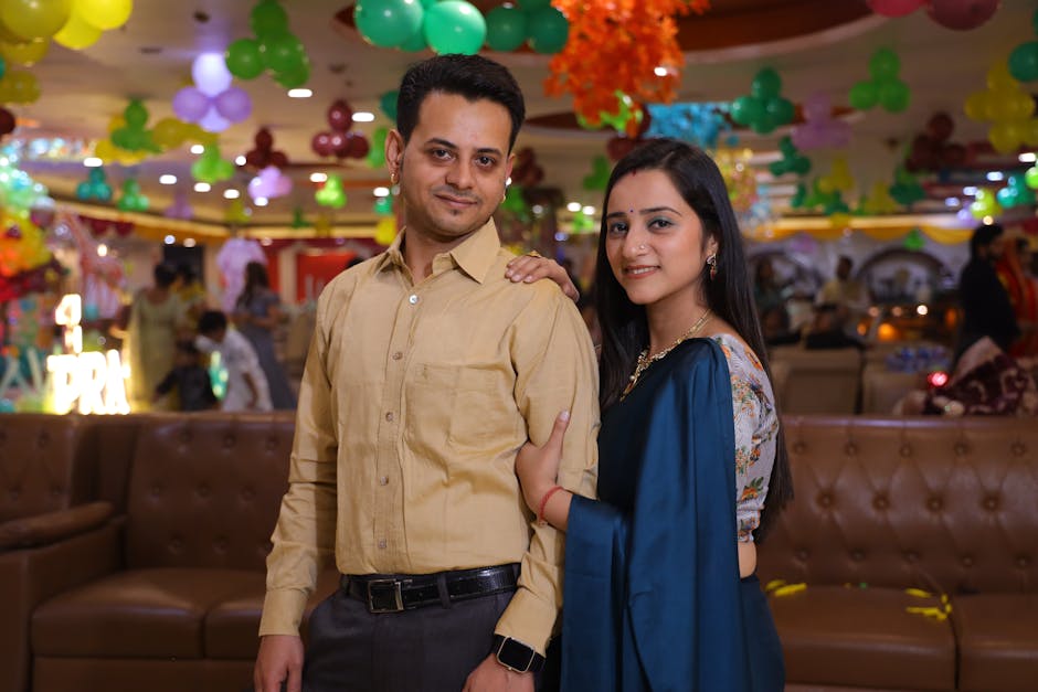 Couple posing happily at an indoor event decorated with colorful balloons and lights.