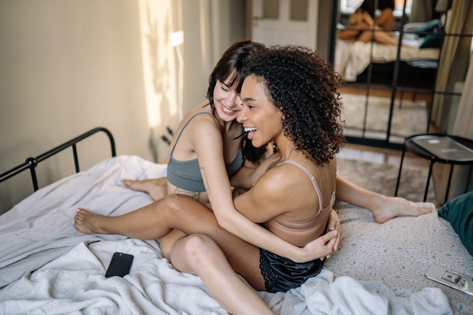 Happy lesbian couple sitting and laughing on bed, embracing love and joy indoors.