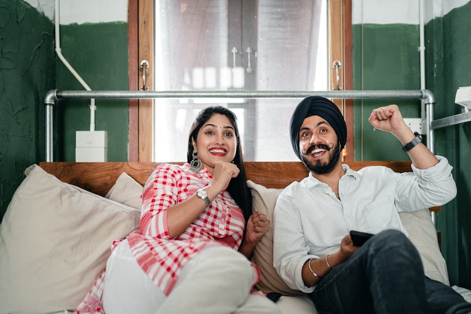 Cheerful Indian couple in casual clothes sitting on sofa and gesticulating hands while watching tv in contemporary living room during weekend