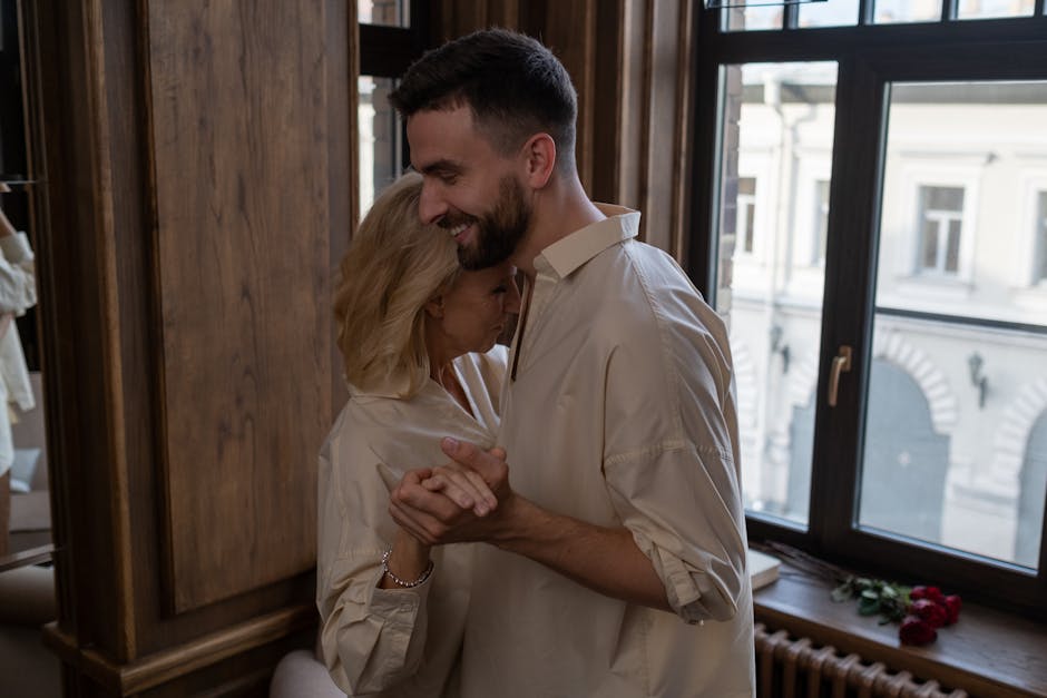 A couple shares a tender dance near a window, bathed in natural light. Captured indoors with love.