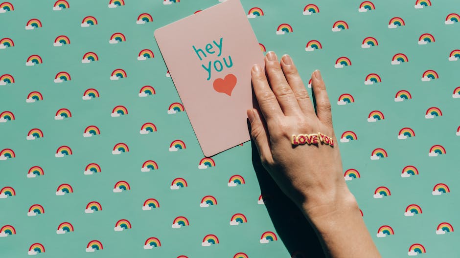 A hand holding a 'hey you' card with a love message on a rainbow patterned background.