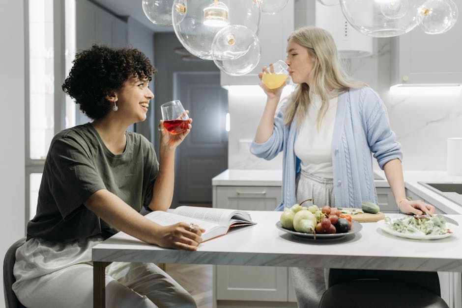 Two women enjoy drinks and conversation around a kitchen table with fruits.