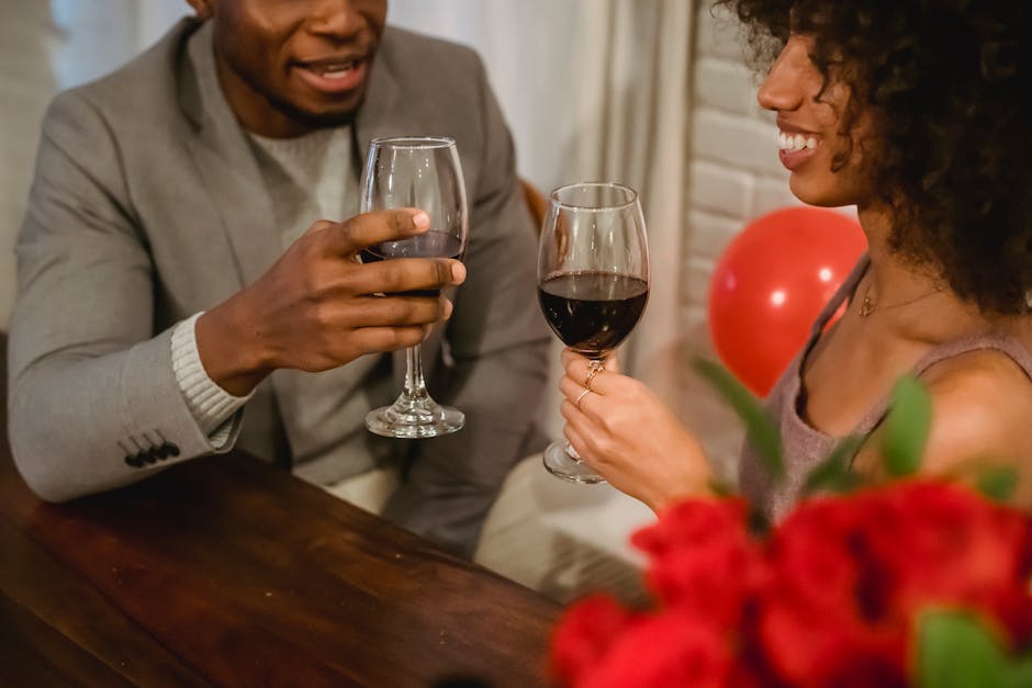 Crop happy young African American couple smiling and clinking wineglasses while celebrating Saint Valentines Day at home decorated with flowers and balloons