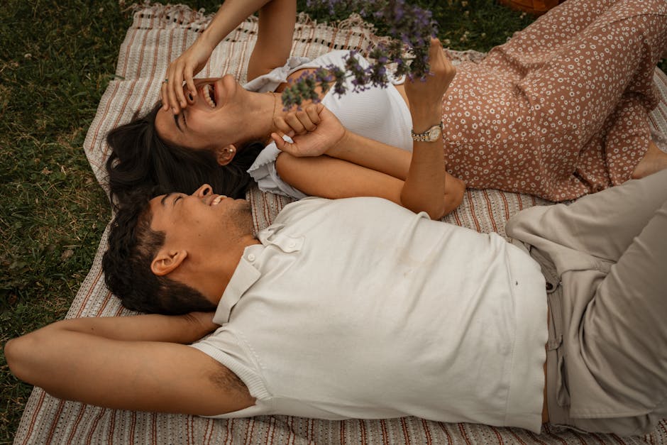 Joyful couple laughing and holding hands, lying on a picnic blanket outdoors.
