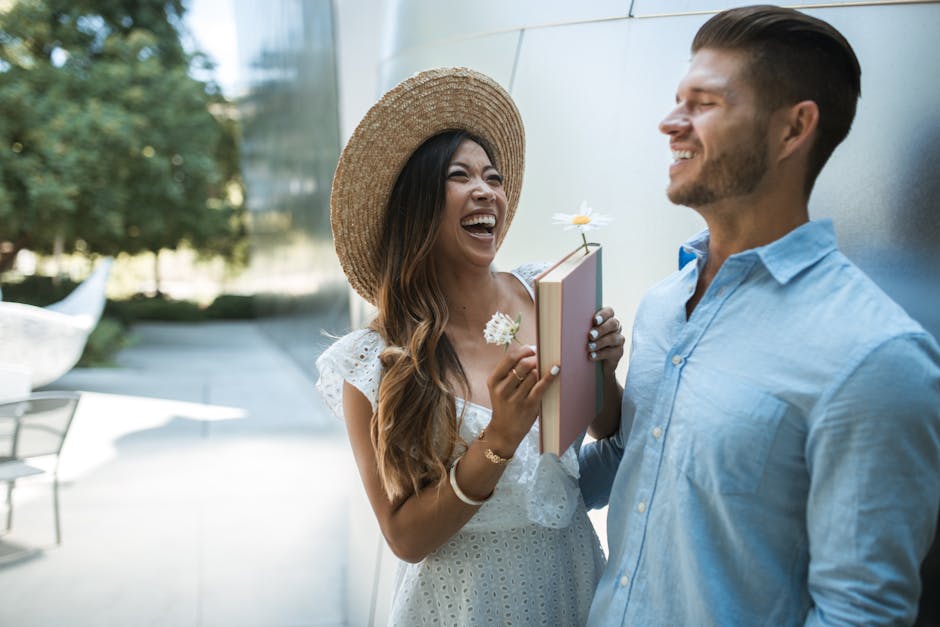 Happy interracial couple laughing while holding a book and flowers outdoors.