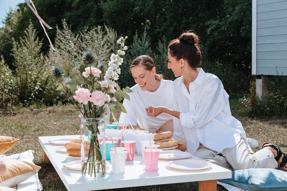 Two women enjoying a sunny outdoor picnic with flowers and snacks.