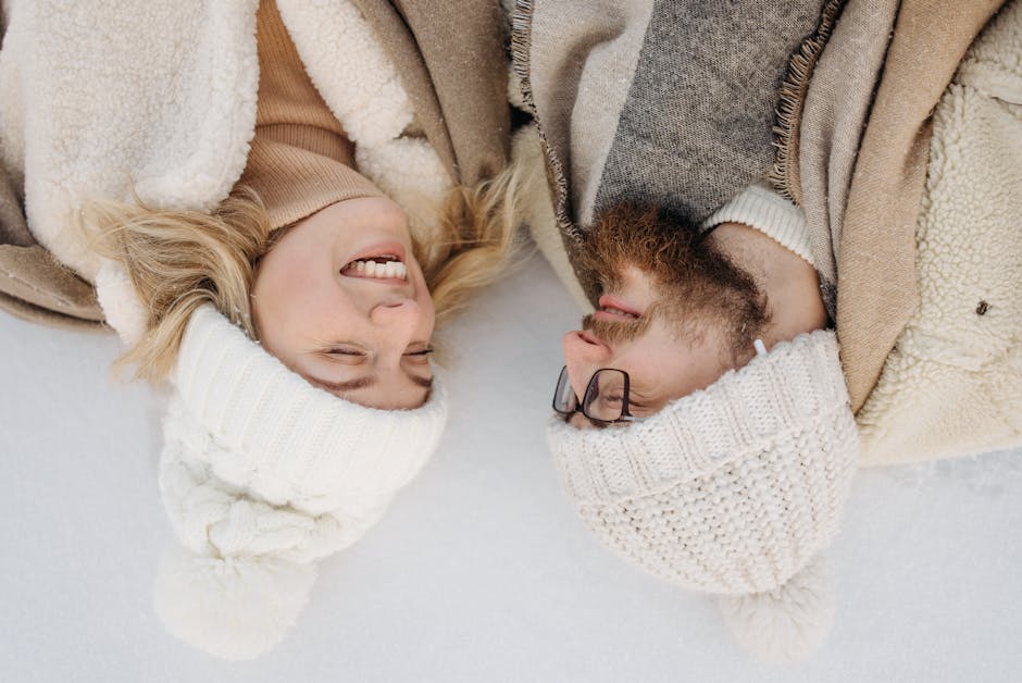 A joyful couple in winter clothing sharing a happy moment lying on the snow, looking at each other during daylight.