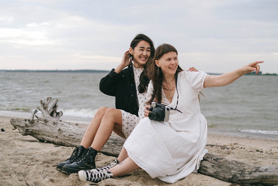 Two smiling women sitting on a beach, capturing memories with a camera.