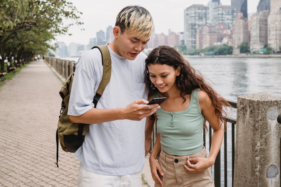 Cheerful young ethnic lady with long hair smiling and watching photos on smartphone with boyfriend while standing on promenade near river in city