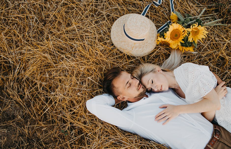 A loving couple rests in a sunny field, embracing amongst sunflowers and straw, expressing tranquility and affection.